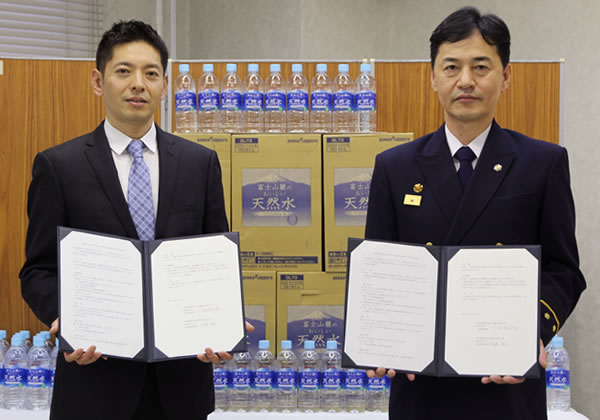 牛込消防署で行われた調印式当日、天然水をバックに協定書を手にする上田署長（当時）と後藤社長（左） 